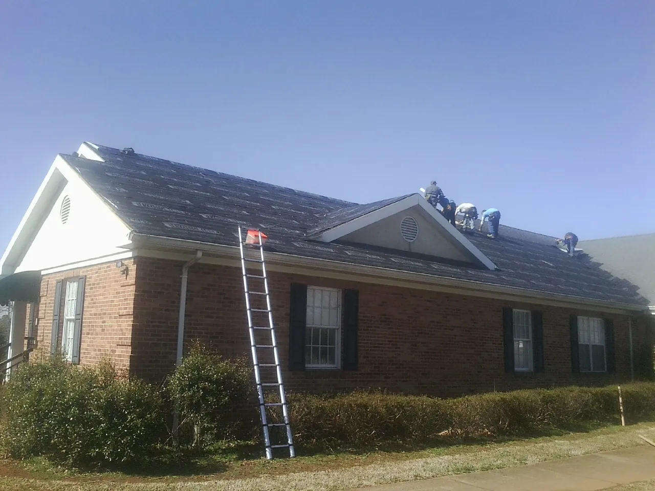 Crew tearing shingles off a brick ranch roof with a ladder set — Emergency Roof Repair work by our crew in Ione