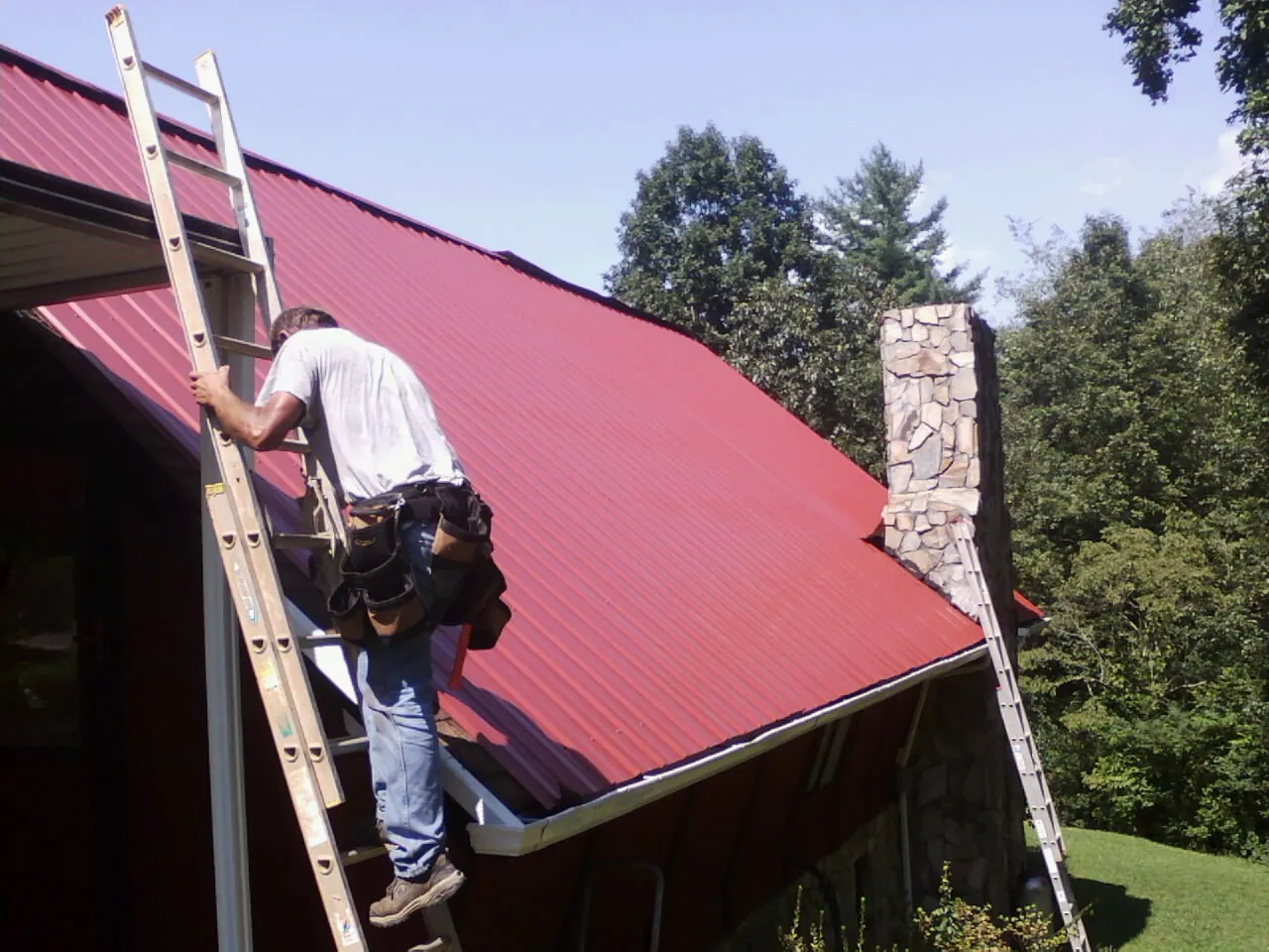 Roofer on a ladder beside a red metal roof and stone chimney — Soffit & Fascia Repair by Roofing Brothers in Ione